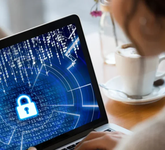 A person seen from over their shoulder is typing on a laptop. The screen displays a cybersecurity graphic with a large, glowing blue padlock icon over streams of digital code.