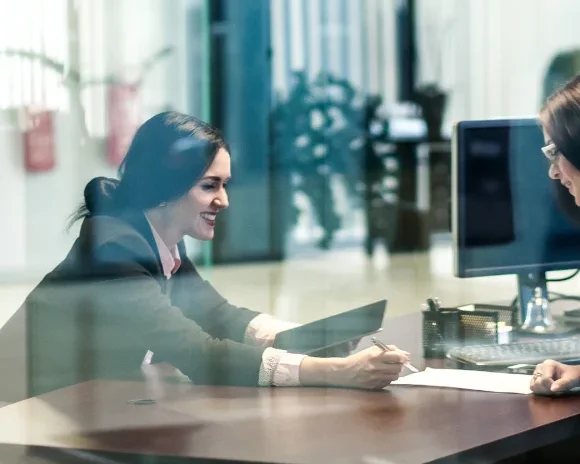 Client-facing side of the cybersecurity consulting industry. In a professional office setting, a consultant sits across the desk from a smiling client. They are reviewing documents together