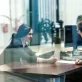 Client-facing side of the cybersecurity consulting industry. In a professional office setting, a consultant sits across the desk from a smiling client. They are reviewing documents together