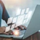 A close-up of a person's hands typing on a laptop, with a bright, warm glow emanating from the keyboard. Floating above the laptop is a transparent digital interface displaying a workflow of glowing document icons.