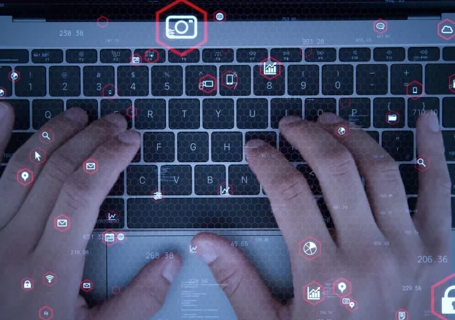 A top-down view of a person's hands typing on a laptop keyboard. A glowing digital overlay with numerous red, hexagonal icons is superimposed on the scene. The icons represent various functions like a camera, a security padlock, cloud storage, and email, conveying a theme of advanced technology, data processing, or cybersecurity.
