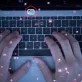 A top-down view of a person's hands typing on a laptop keyboard. A glowing digital overlay with numerous red, hexagonal icons is superimposed on the scene. The icons represent various functions like a camera, a security padlock, cloud storage, and email, conveying a theme of advanced technology, data processing, or cybersecurity.