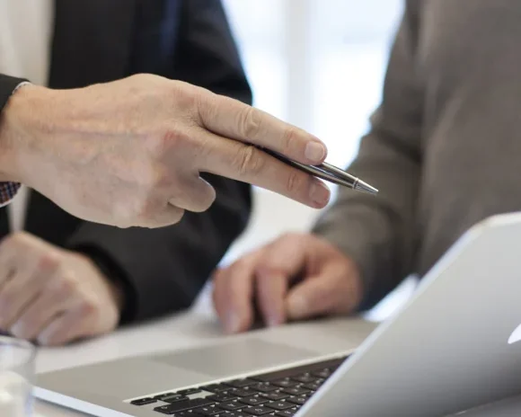 Two IT professionals discussing information displayed on a laptop screen, with one person pointing at the screen using a pen during a collaborative meeting.