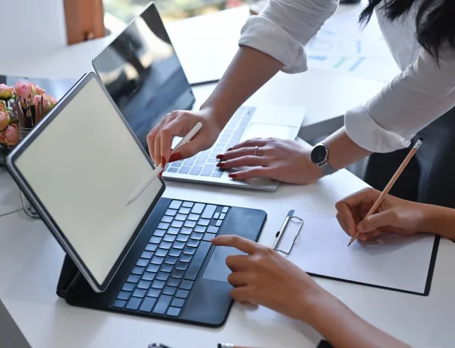 A close-up of a collaborative workspace where two people are working together at a white desk. One person stands and points at a laptop screen, while the other person sits, pointing at the screen of a tablet computer while holding a pencil over a notepad on a clipboard.