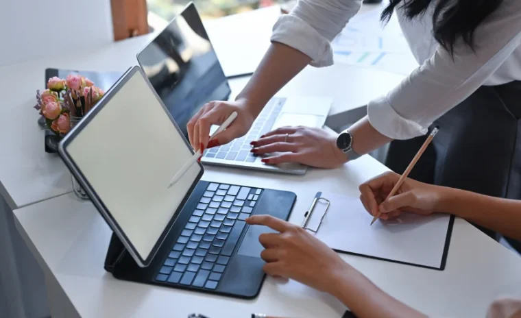 A close-up of a collaborative workspace where two people are working together at a white desk. One person stands and points at a laptop screen, while the other person sits, pointing at the screen of a tablet computer while holding a pencil over a notepad on a clipboard.