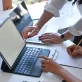 A close-up of a collaborative workspace where two people are working together at a white desk. One person stands and points at a laptop screen, while the other person sits, pointing at the screen of a tablet computer while holding a pencil over a notepad on a clipboard.