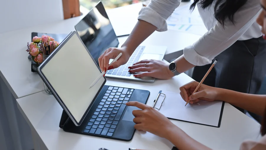 A close-up of a collaborative workspace where two people are working together at a white desk. One person stands and points at a laptop screen, while the other person sits, pointing at the screen of a tablet computer while holding a pencil over a notepad on a clipboard.