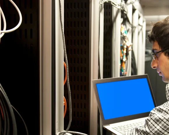IT technician working in a server room, connecting network cables while using a laptop with a blue screen for diagnostics.