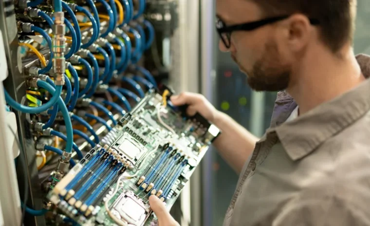 An IT technician with glasses and a beard is shown from the side, carefully installing a large computer motherboard into a server rack. The background is a dense network of bright blue and yellow cables, indicative of a data center environment.