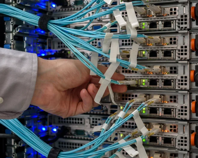 A close-up of a network technician's hand organizing a bundle of neatly labeled, bright blue fiber optic cables in a server rack. The cables are plugged into network switches, which have glowing green and blue indicator lights showing network activity.